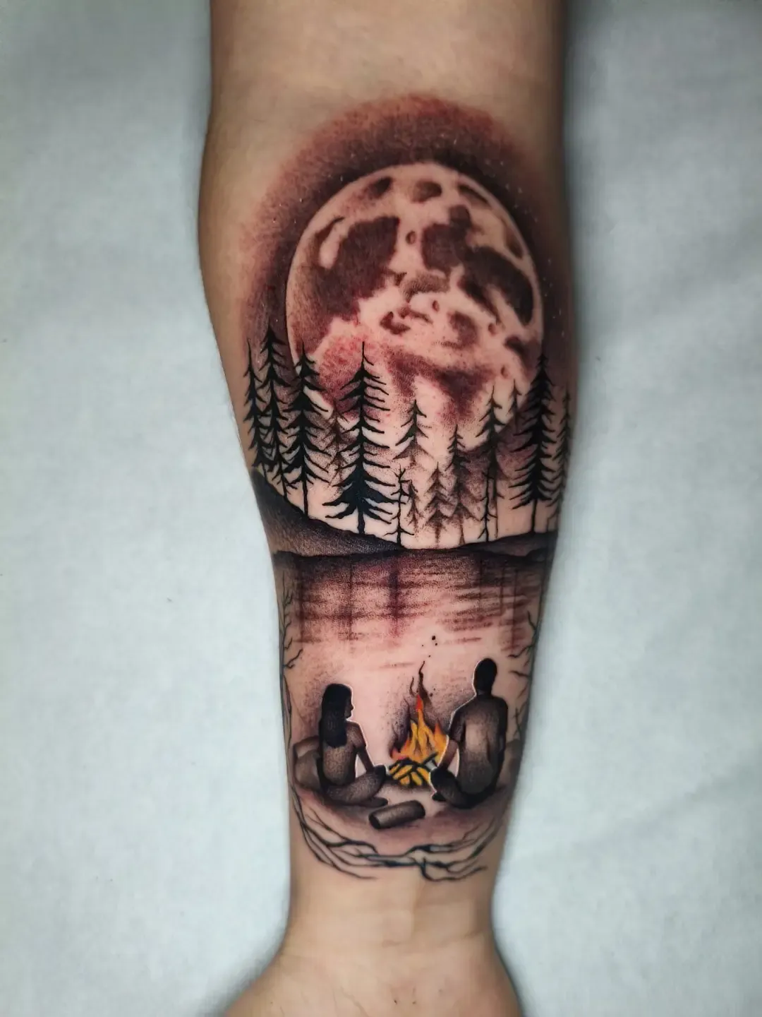 Tatuaje ilustrativo de luna llena, bosque y hoguera en el antebrazo